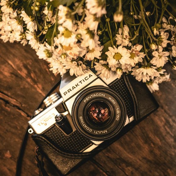 A vintage Praktica camera surrounded by delicate white flowers on a wooden table.