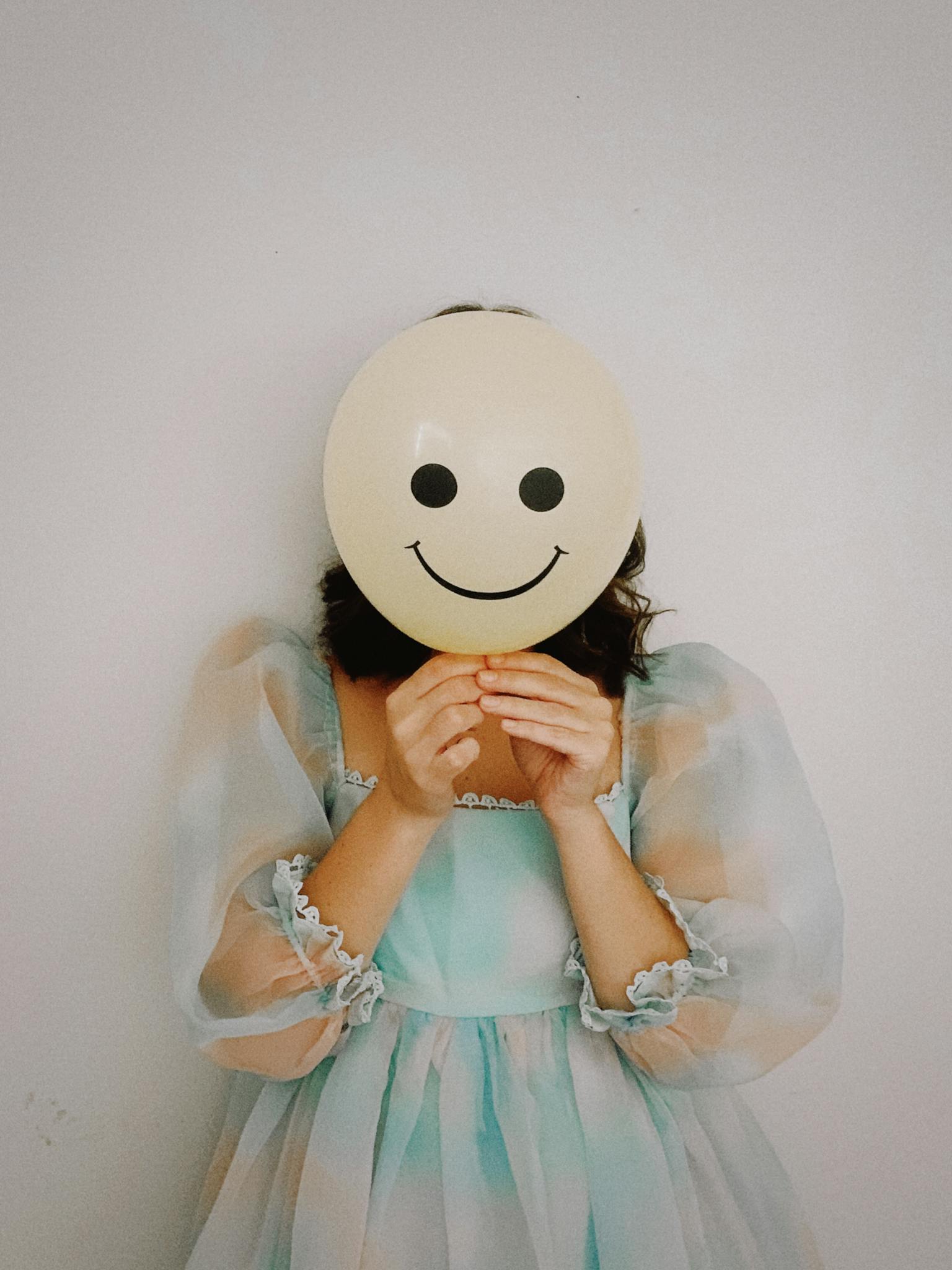 Elegant woman in a pastel dress holding a smiley face balloon against a simple background.
